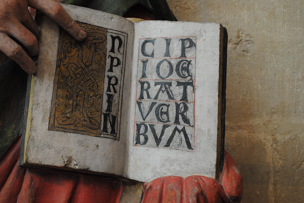 Ge&ouml;ffnetes Buch mit den Anfangsworten des Johannes-Evangeliums IN PRINCIPIO ERAT VERBVM in den H&auml;nden einer Steinfigur des Evangelisten (um 1260), Dom, Mei&szlig;en. Die gemalte Doppelseite, die an Evangelien-Anf&auml;nge in mittelalterlichen Handschriften erinnert, weist mehrere Farbfassungen auf. W&auml;hrend die goldene Schmuckinitiale noch zur Herstellungszeit der Figur geh&ouml;rt, entstanden die schwarzen Majuskelbuchstaben erst um 1500 durch &Uuml;bermalen der urspr&uuml;nglichen Inschrift.