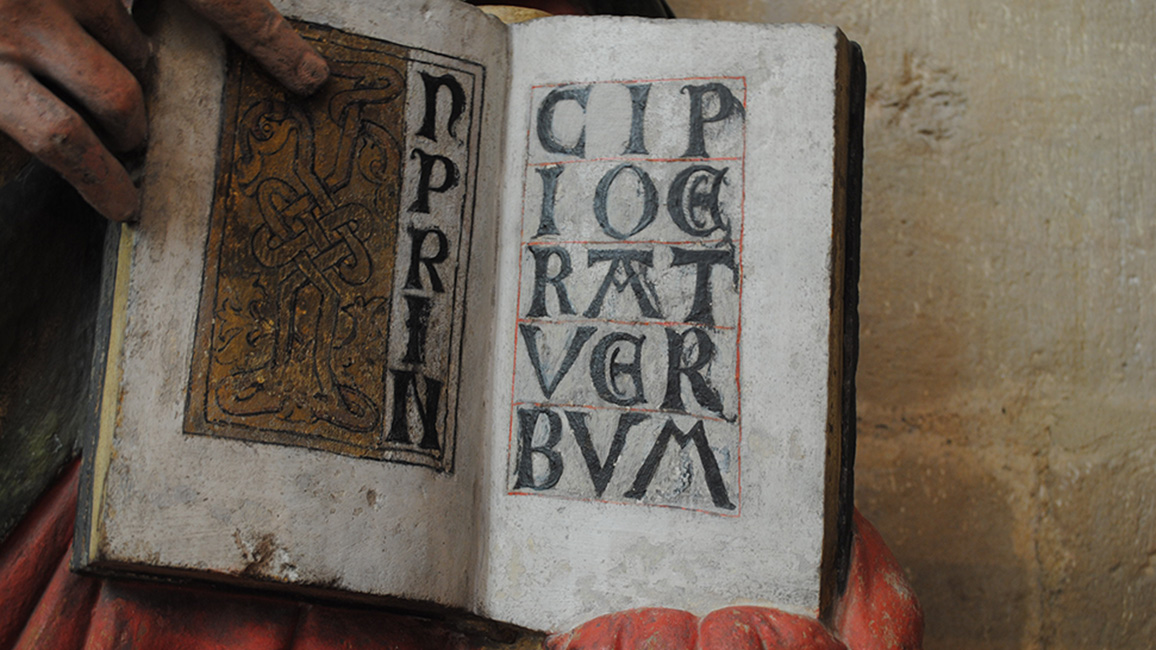 Ge&ouml;ffnetes Buch mit den Anfangsworten des Johannes-Evangeliums IN PRINCIPIO ERAT VERBVM in den H&auml;nden einer Steinfigur des Evangelisten (um 1260), Dom, Mei&szlig;en. Die gemalte Doppelseite, die an Evangelien-Anf&auml;nge in mittelalterlichen Handschriften erinnert, weist mehrere Farbfassungen auf. W&auml;hrend die goldene Schmuckinitiale noch zur Herstellungszeit der Figur geh&ouml;rt, entstanden die schwarzen Majuskelbuchstaben erst um 1500 durch &Uuml;bermalen der urspr&uuml;nglichen Inschrift.