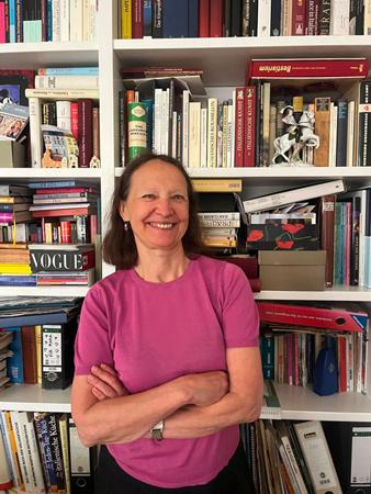 Frau mit kurzen braunen Haaren und pinkem T-Shirt steht vor einem vollgestellten B&uuml;cherregal und l&auml;chelt mit verschr&auml;nkten Armen